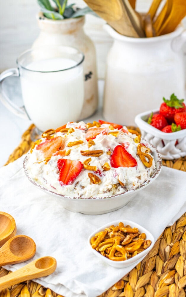 A bowl of creamy dessert topped with sliced strawberries and pretzels, with a glass of milk, fresh strawberries, and wooden utensils in the background.