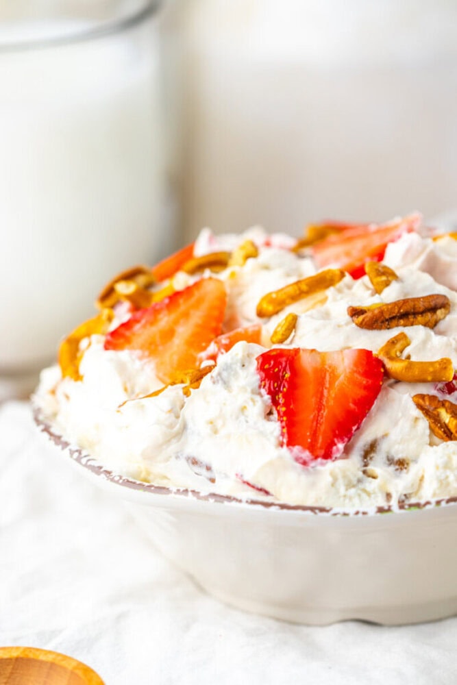 A bowl of creamy dessert topped with sliced strawberries and pretzel pieces, with a glass of milk in the background.
