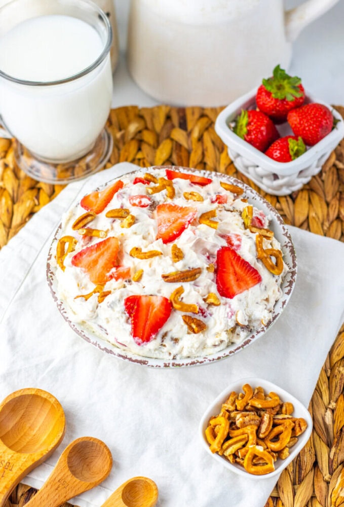 A bowl of creamy dessert with sliced strawberries and pretzel pieces, surrounded by a glass of milk, a dish of strawberries, wooden spoons, and a small bowl of pretzels.