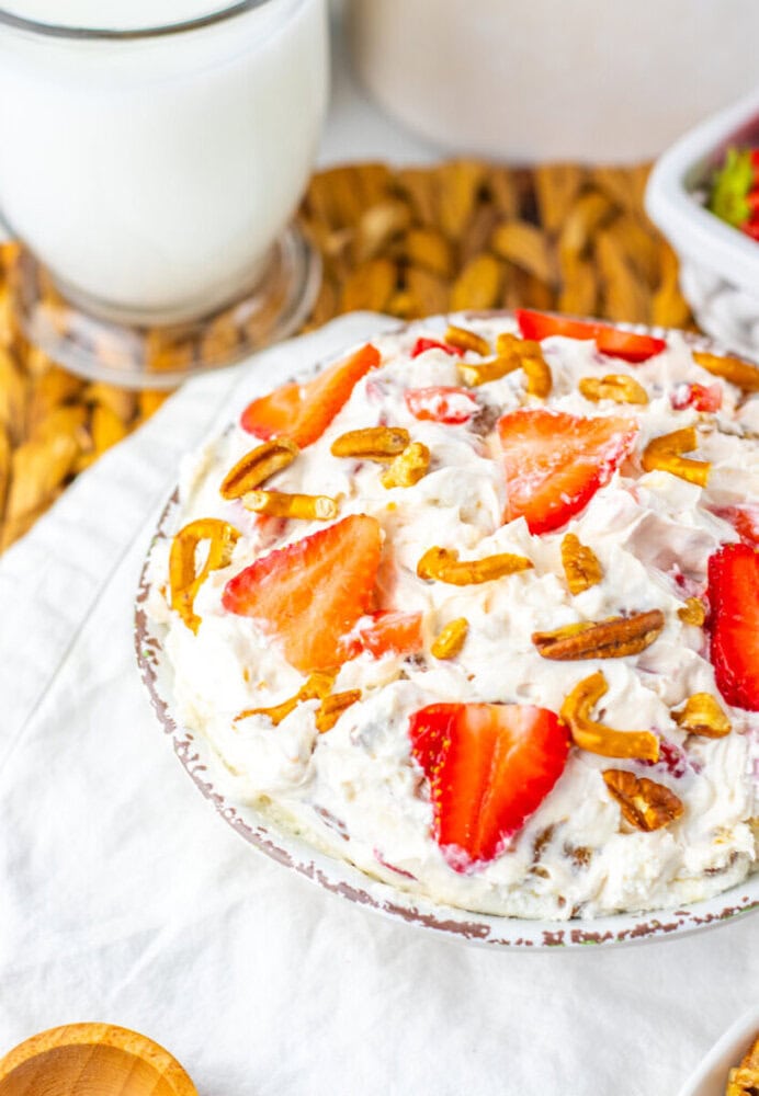 A bowl of creamy dessert topped with sliced strawberries and pretzel pieces, placed on a white cloth with a glass of milk in the background.