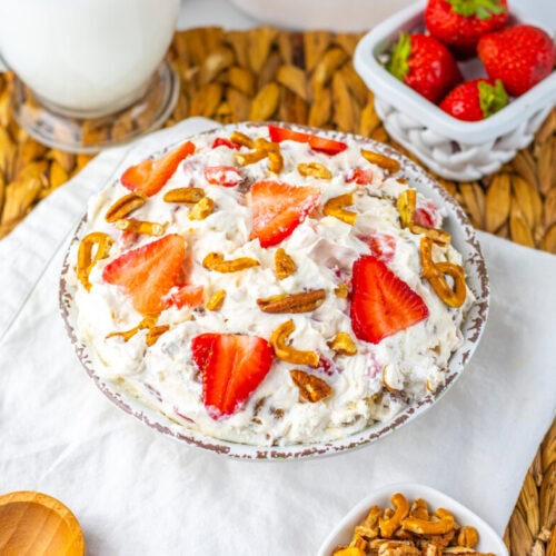 A bowl of creamy salad topped with sliced strawberries and pretzel pieces, placed on a white napkin with a small dish of strawberries and a glass of milk nearby.