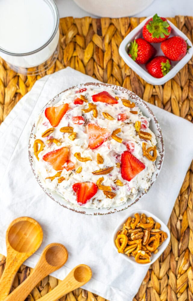 A bowl of strawberry pretzel salad topped with sliced strawberries and pretzel pieces, surrounded by a glass of milk, strawberries, small pretzel pieces, and wooden measuring spoons.
