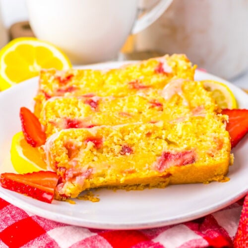 Three slices of strawberry lemon cake are arranged on a white plate with fresh strawberry and lemon slices, placed on a red and white checkered cloth.