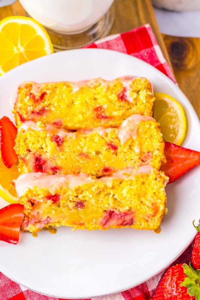 Three slices of lemon strawberry bread with glaze are arranged on a white plate, garnished with fresh lemon slices and strawberries.
