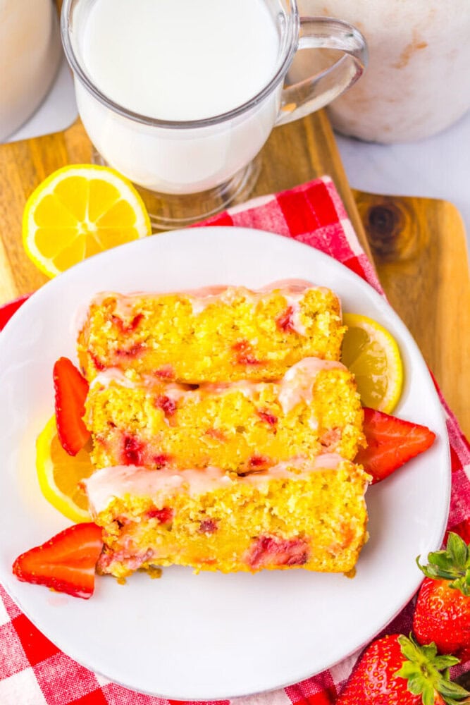 A plate with three slices of strawberry lemon bread, garnished with strawberry and lemon slices, next to a glass mug of milk on a red checked napkin.