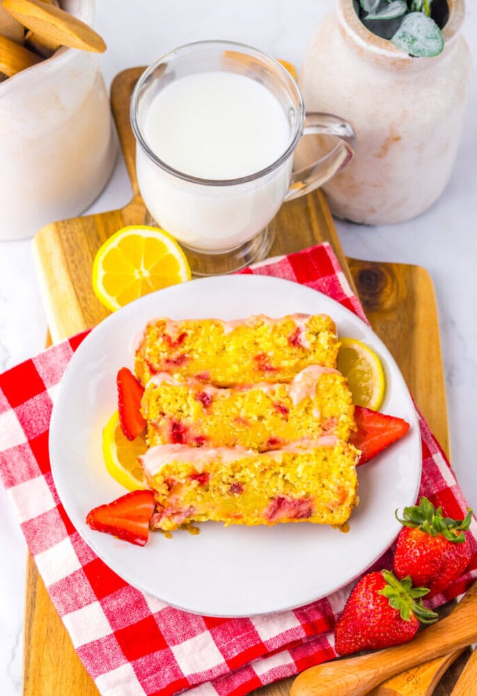 A plate with three slices of lemon strawberry bread garnished with fresh strawberries and lemon slices, next to a glass of milk on a wooden board with a red checkered napkin.