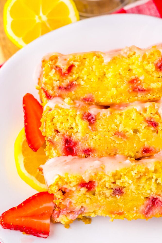 Three slices of lemon strawberry loaf cake with glaze are served on a white plate, garnished with fresh lemon slices and strawberries.