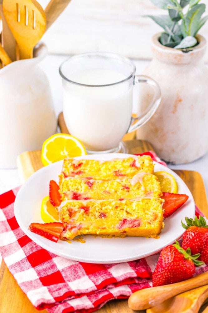 Three slices of lemon strawberry bread are served on a white plate with fresh strawberries. A glass of milk, lemon slices, and kitchen utensils are in the background.