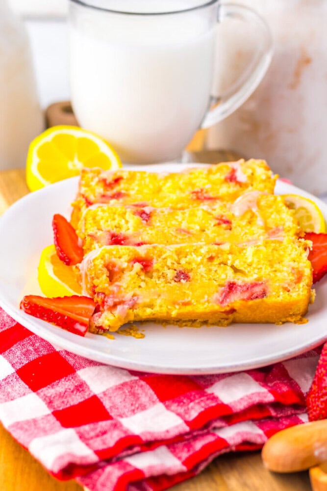 Three slices of lemon strawberry bread on a white plate, garnished with fresh strawberries and lemon slices, on a red checkered napkin with a glass of milk in the background.