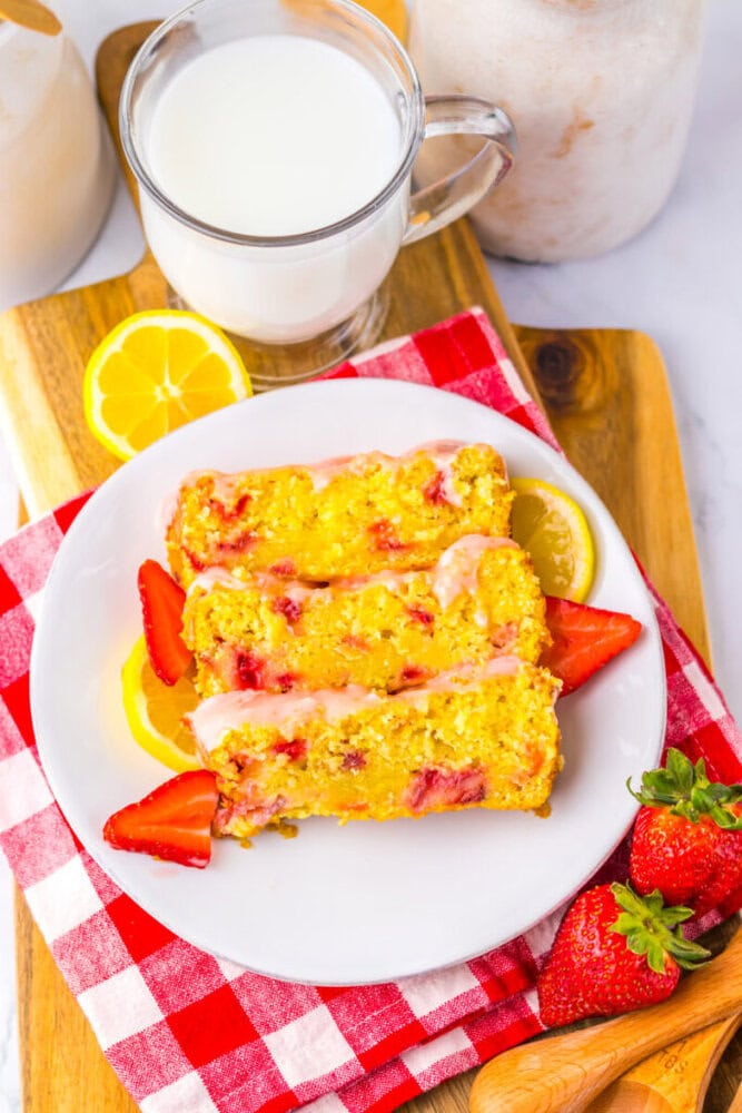 Three slices of strawberry lemon bread on a white plate with fresh strawberries and lemon slices, next to a glass of milk on a red checkered napkin.