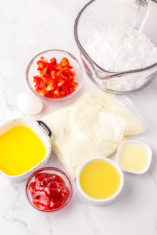 Assorted baking ingredients on a white surface, including chopped strawberries, powdered sugar, an egg, melted butter, cake mix, strawberry jam, lemon juice, and another liquid in small bowls.
