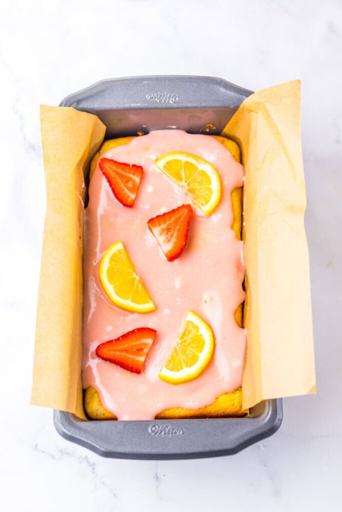 A loaf cake in a lined pan topped with pink icing, lemon slices, and strawberry pieces on a white marble surface.
