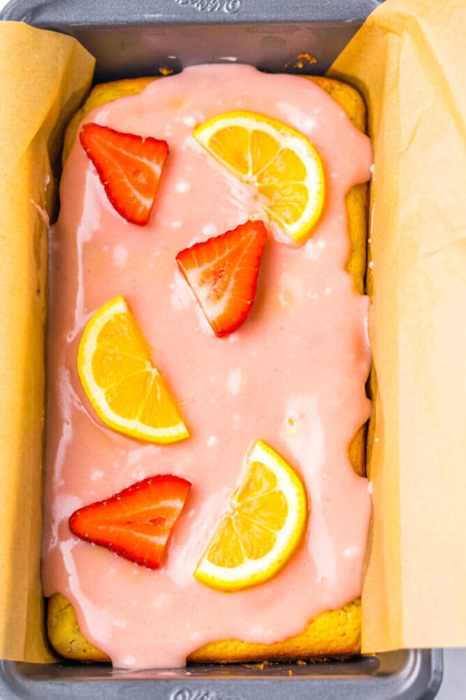 A rectangular loaf cake with pink icing, topped with lemon slices and halved strawberries, sits in a parchment-lined baking pan.