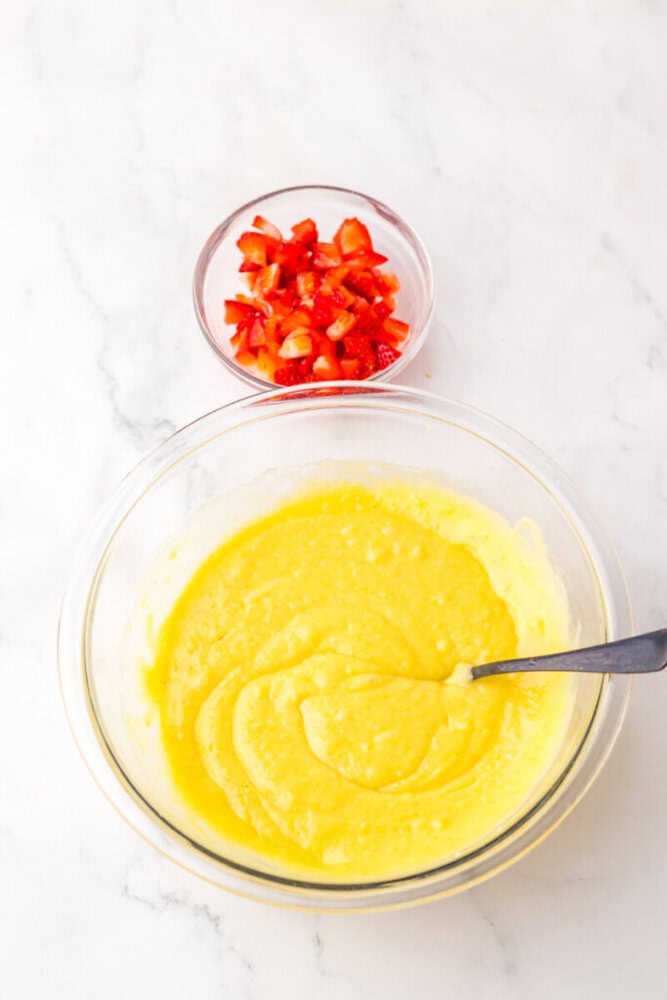 A glass bowl with yellow cake batter and a spoon, next to a small bowl of chopped strawberries on a white surface.