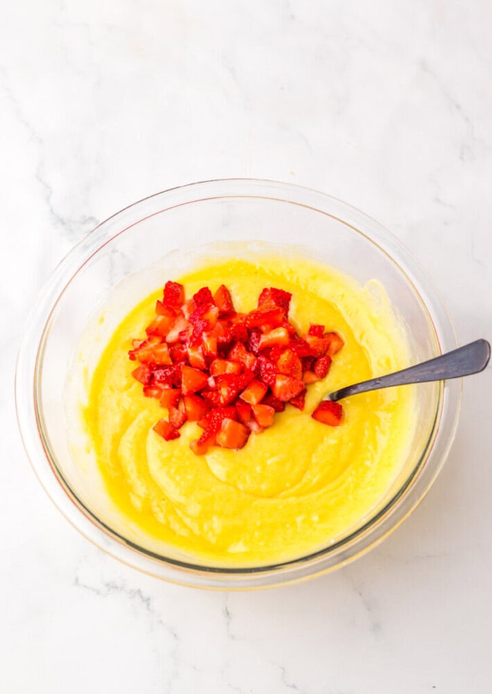 A glass bowl filled with yellow batter and topped with chopped strawberries, with a spoon resting inside, on a white marble surface.