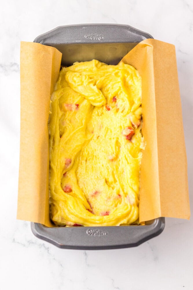 A parchment-lined loaf pan filled with yellow cake batter containing small pieces of fruit, ready to be baked.
