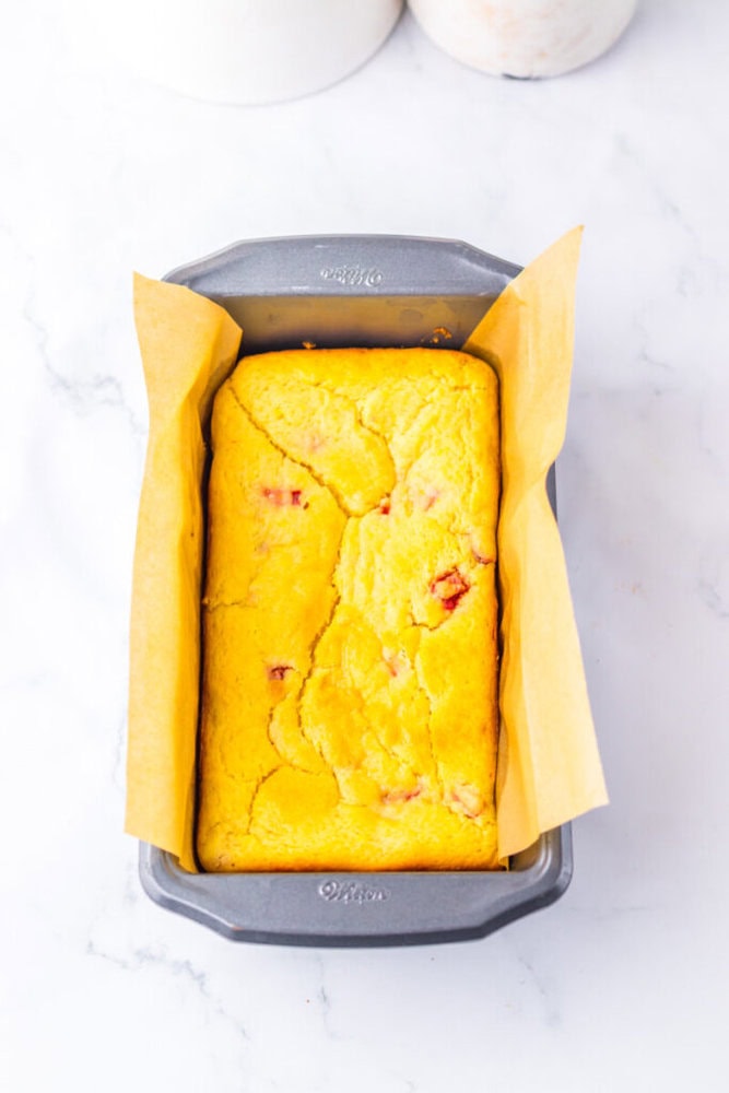 A rectangular loaf of cornbread with visible pieces of red pepper sits in a parchment-lined metal baking pan on a white surface.