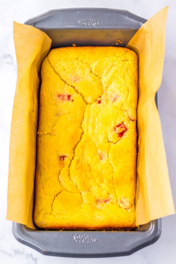 A rectangular loaf of cornbread with visible cracks on top, baked in a parchment-lined metal pan.