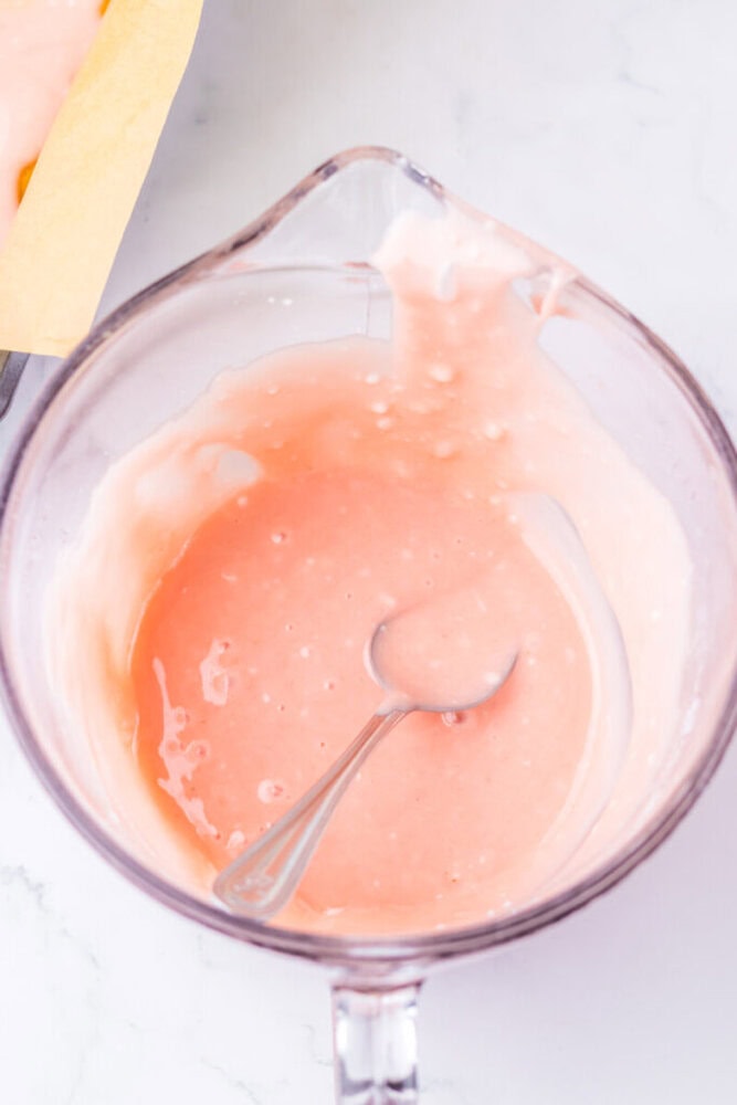 A glass mixing bowl with pink batter and a metal spoon inside, sitting on a white countertop.