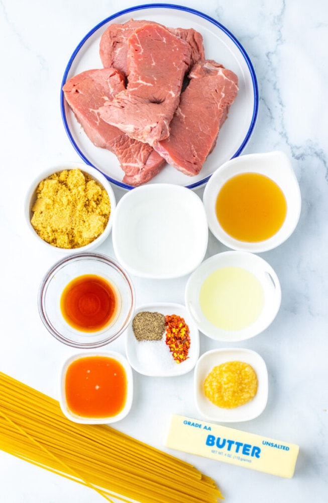 A collection of ingredients on a white surface, including raw steak, uncooked spaghetti, butter, seasonings, liquid sauces, grated cheese, and small bowls of various liquids.