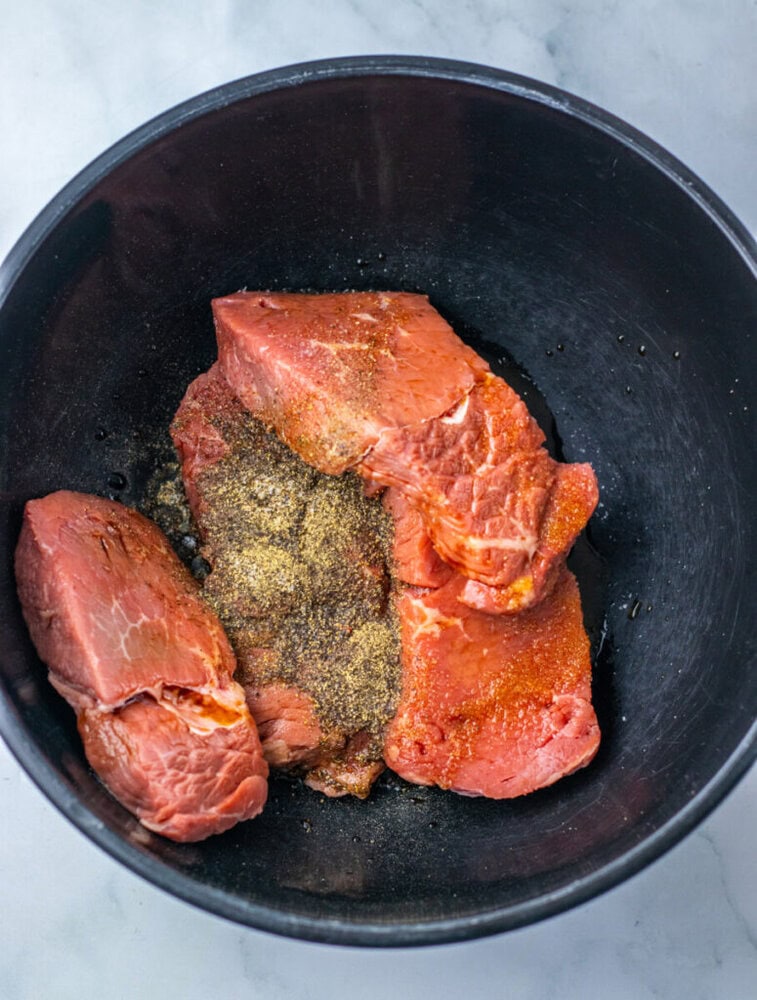 Raw beef pieces seasoned with ground black pepper and spices in a black mixing bowl on a light-colored surface.