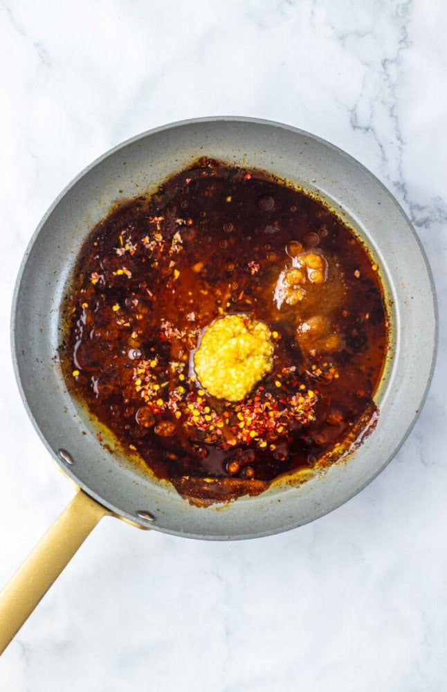 A frying pan containing a dark sauce with visible red pepper flakes and a dollop of minced garlic sits on a white marble surface.