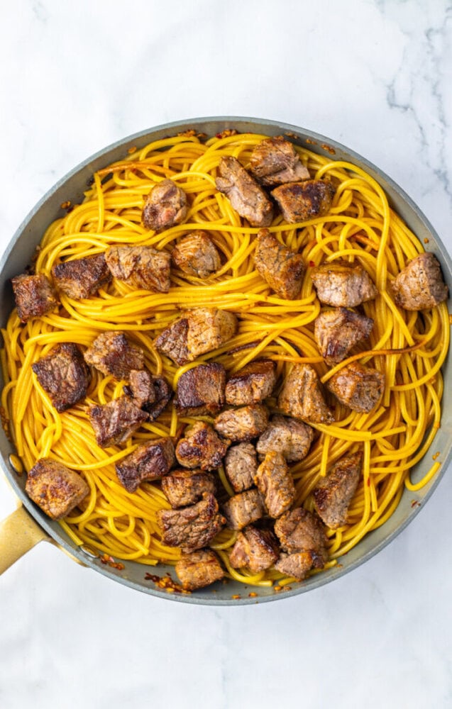 Cooked spaghetti and browned steak cubes in a skillet on a marble surface.