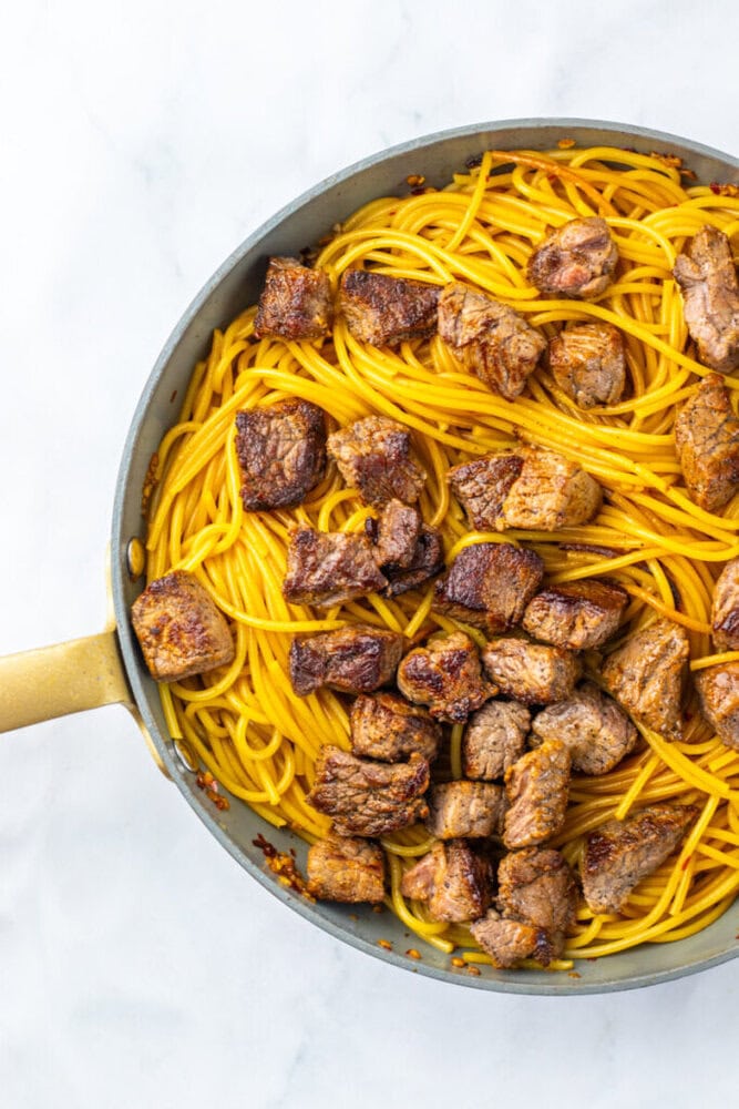 A skillet filled with cooked spaghetti and browned beef cubes on a white surface.