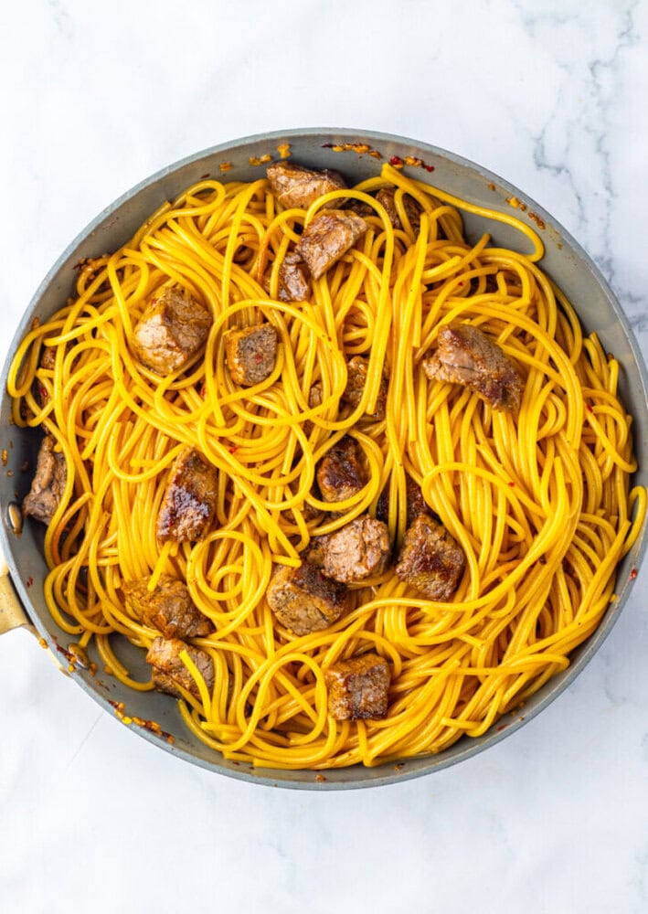 A pan filled with cooked spaghetti and seared pieces of beef, all lightly coated in a yellow-orange sauce, on a white marble surface.