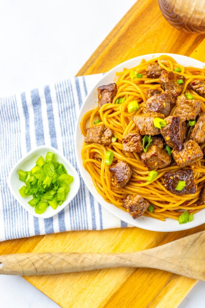 A plate of spaghetti with chunks of beef and chopped green onions, served beside a small dish of green onions on a striped napkin.