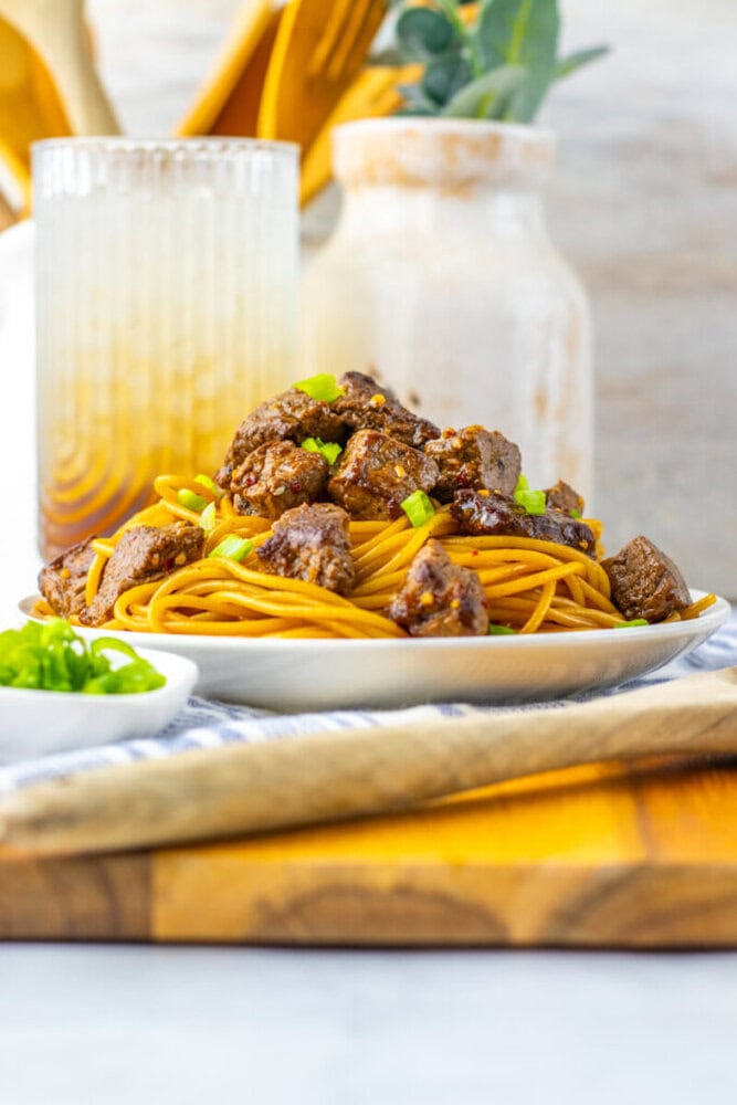 A plate of spaghetti topped with chunks of beef and green onions sits on a table with a glass of drink and a bowl of sliced green onions in the background.