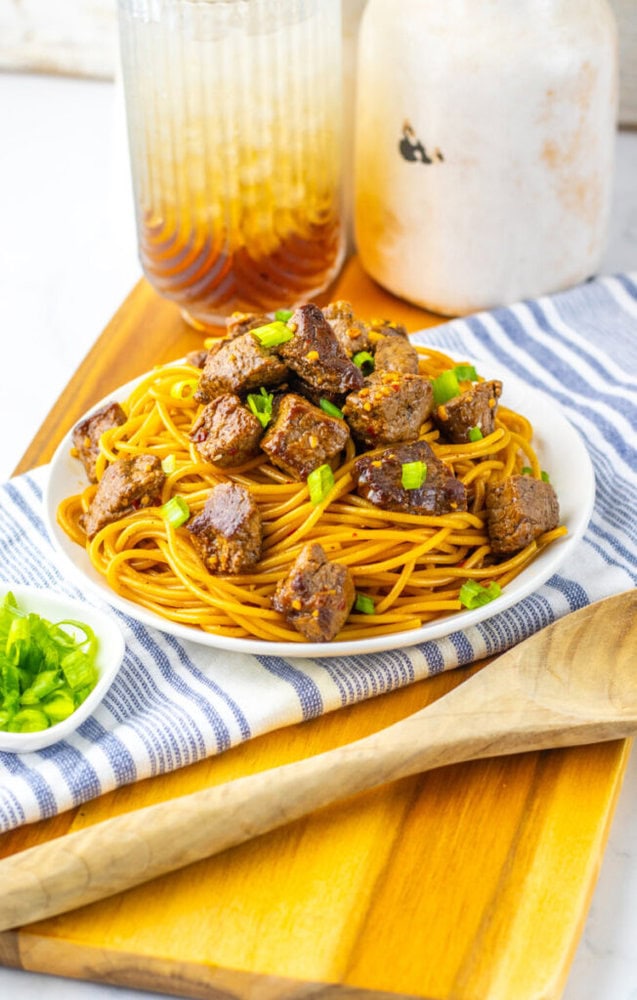 A plate of spaghetti topped with cubed beef and chopped green onions sits on a wooden board next to a wooden spoon and a pitcher of iced tea.