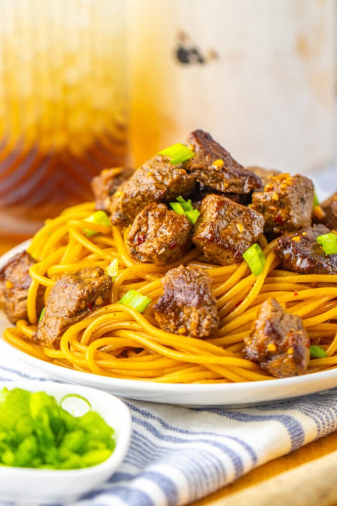A plate of cooked spaghetti topped with seasoned beef cubes and garnished with chopped green onions, with a small serving of sliced green vegetables on the side.