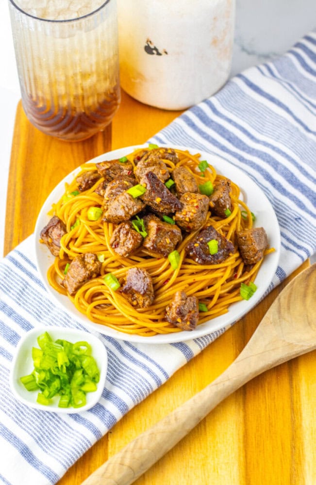 A plate of spaghetti with beef cubes and chopped green onions, served on a striped napkin with a wooden spoon, a small bowl of green onions, and a glass of iced drink nearby.