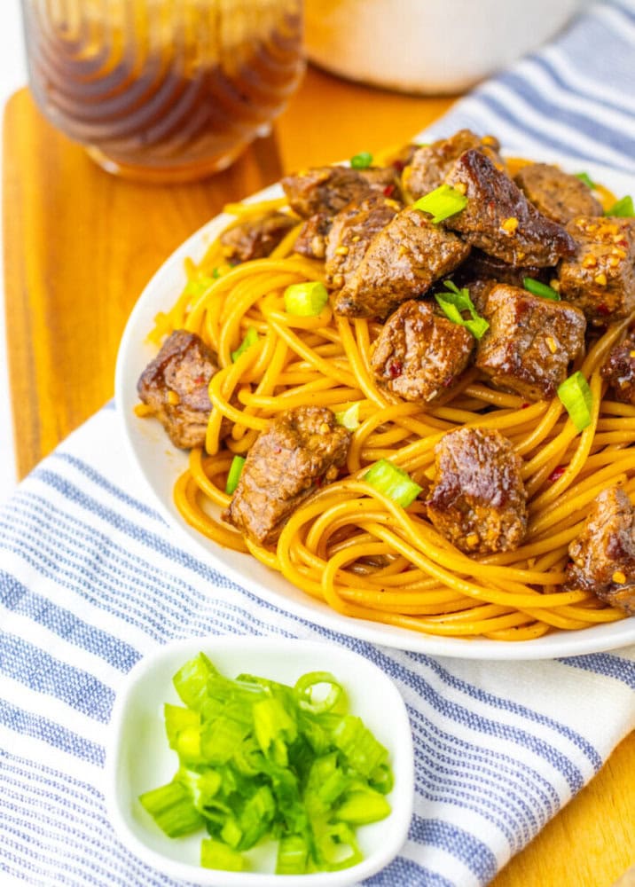 A plate of spaghetti topped with seared beef cubes and chopped green onions, accompanied by a small bowl of sliced green onions and a glass of iced beverage in the background.