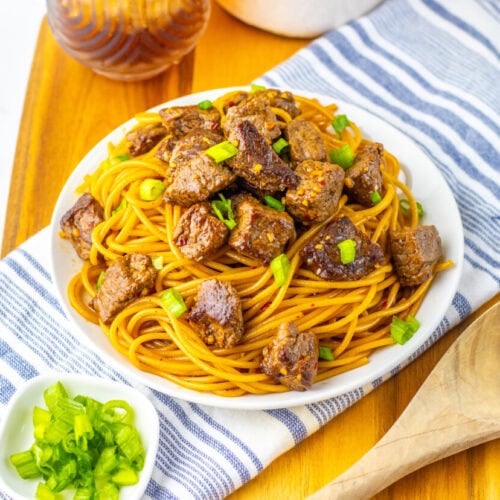 A plate of spaghetti topped with chunks of cooked meat and chopped green onions, placed on a striped cloth with a wooden spoon and a small bowl of green onions nearby.