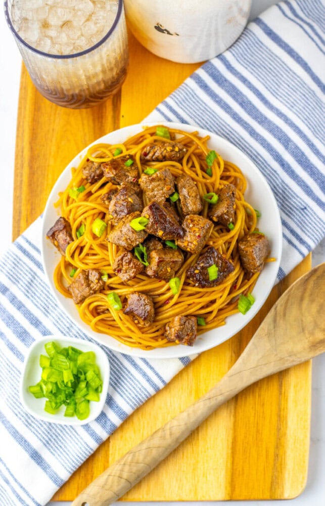 A plate of noodles topped with cubed beef and chopped green onions, placed on a striped napkin with a glass of iced drink and a wooden spoon nearby.