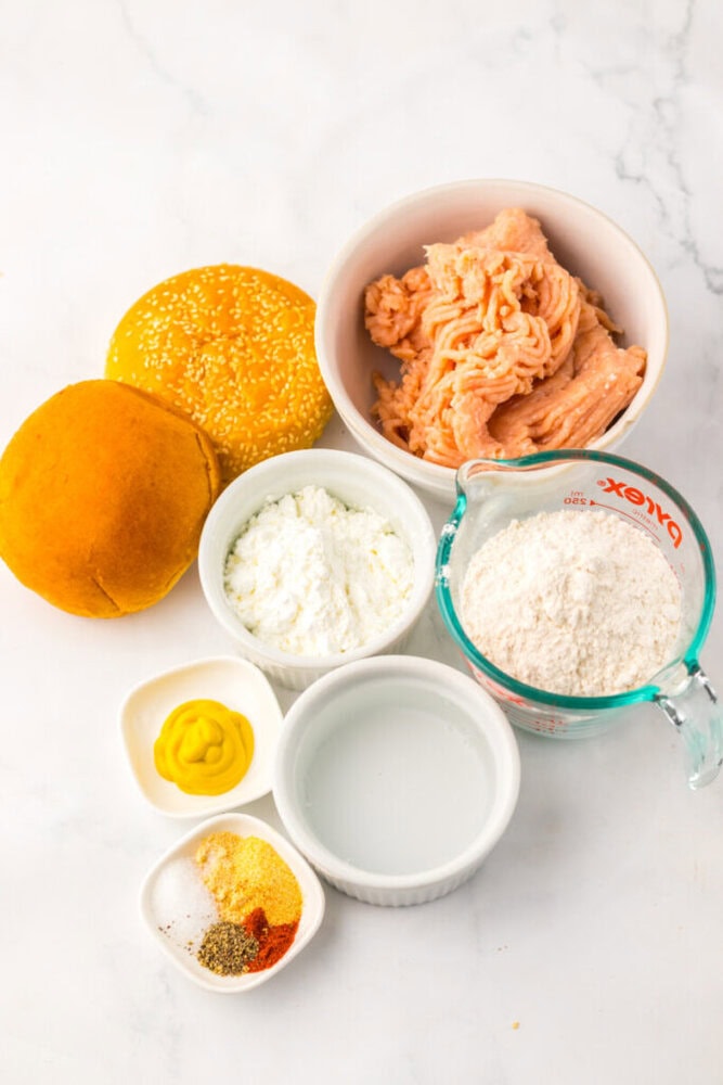 Bowls containing ground chicken, flour, cornstarch, spices, mustard, water, and two sandwich buns are arranged on a white surface.