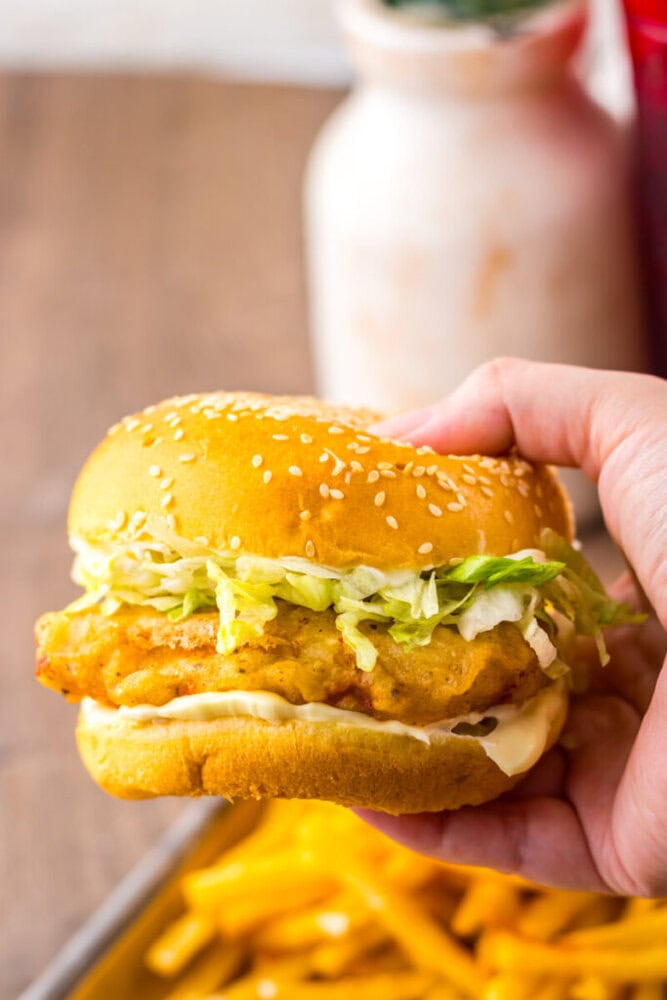 A hand holding a chicken sandwich with shredded lettuce and mayonnaise in a sesame seed bun, with a tray of fries in the background.