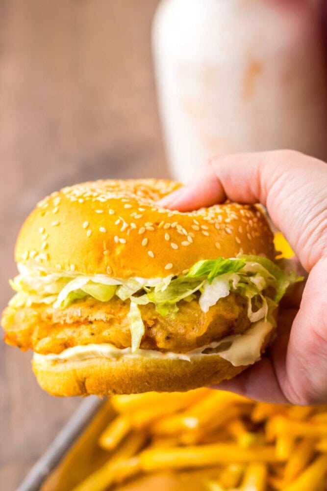 A hand holding a sesame seed chicken sandwich with lettuce and mayonnaise, with a tray of fries and a blurred drink in the background.