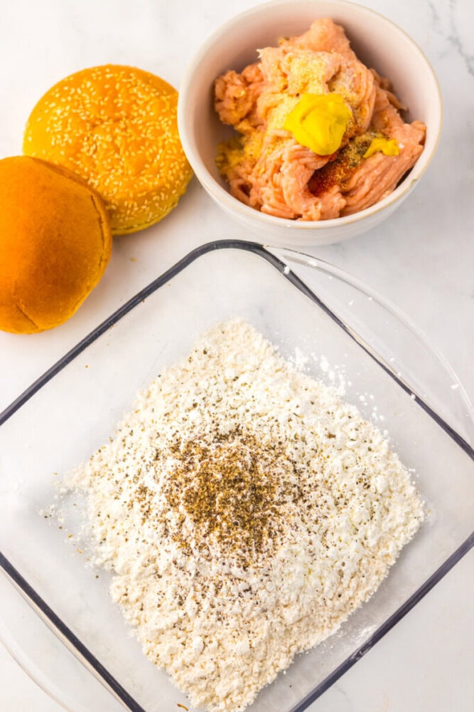 Raw ground chicken with seasoning in a bowl, burger buns beside it, and a dish of flour and spices on a marble surface.