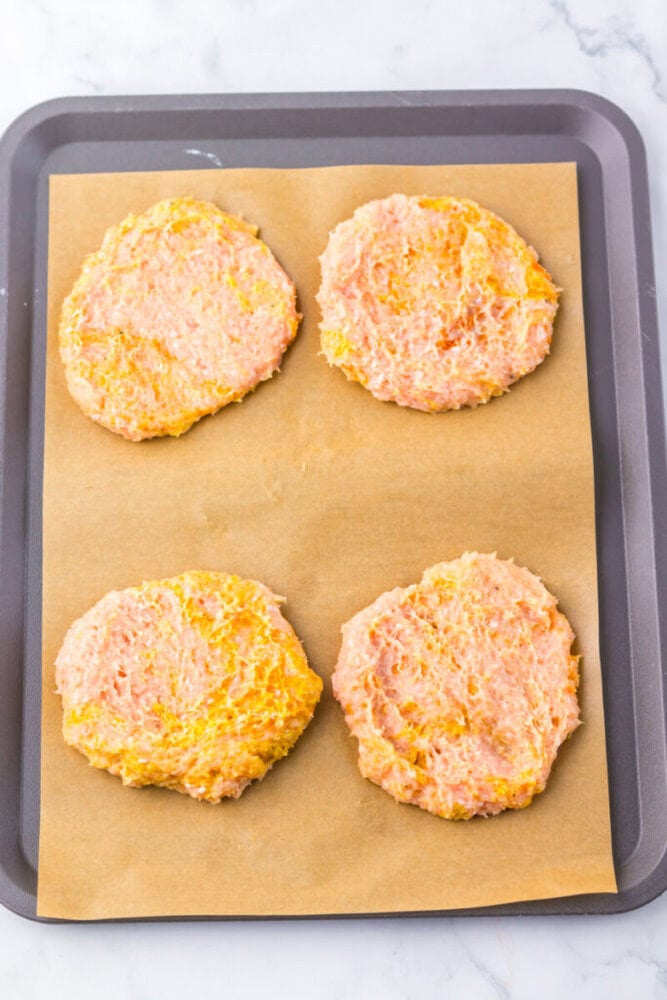 Four raw chicken patties are placed on a baking tray lined with parchment paper, ready to be cooked.