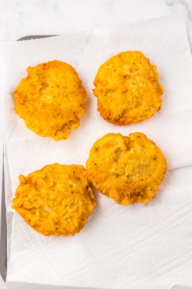 Four golden-brown fritters rest on a white paper towel, which is absorbing excess oil on a flat surface.