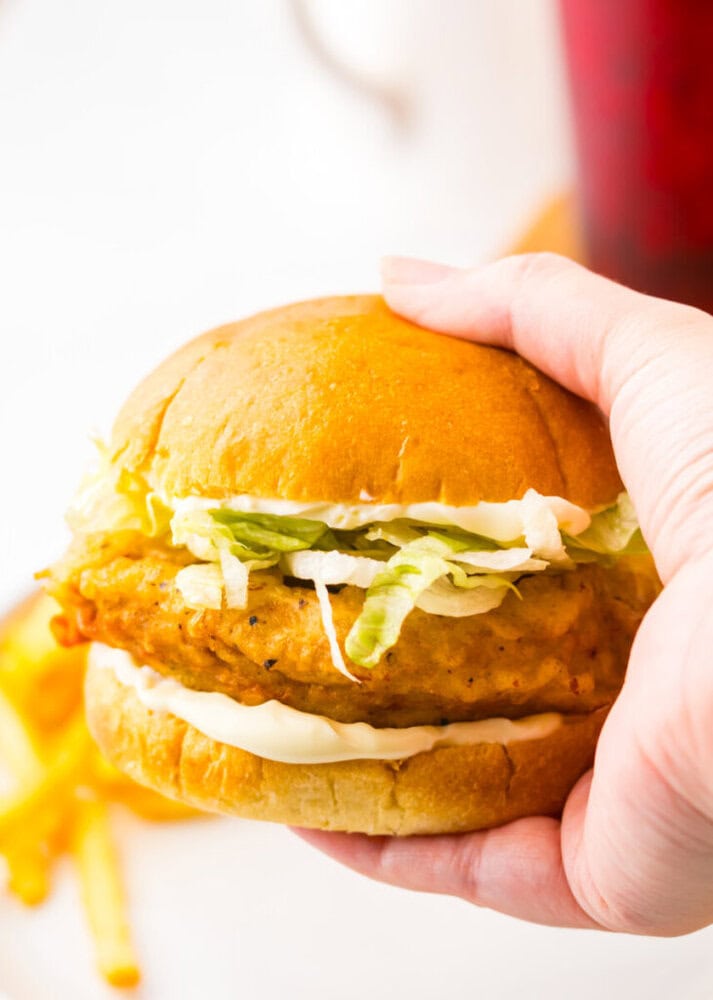 A hand is holding a chicken sandwich with lettuce and mayonnaise in a bun. French fries are visible in the background.