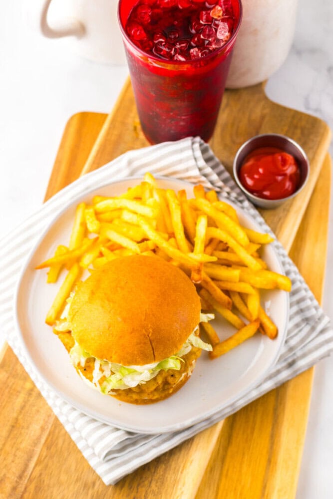 A plate with a sandwich, French fries, and a side of ketchup, alongside a red iced drink on a wooden board with a striped napkin.