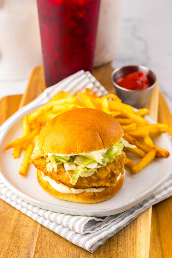 A chicken sandwich with lettuce and sauce on a bun is served with French fries and a cup of ketchup on a white plate, with a red drink in the background.