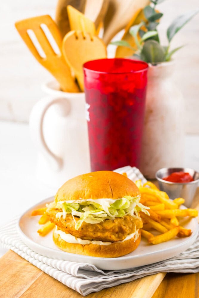 A fried chicken sandwich with lettuce and mayonnaise is served on a plate with French fries. A red cup, ketchup, and kitchen utensils are in the background.