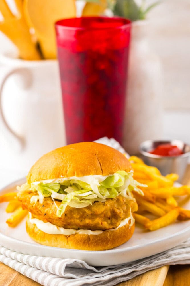 A fried chicken sandwich with lettuce and mayonnaise is served with French fries and a small cup of ketchup, next to a red drink.