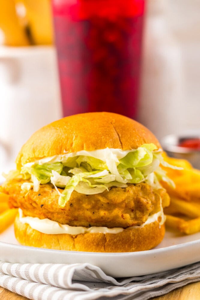 A crispy chicken sandwich with lettuce and mayonnaise on a bun, served with French fries and a red beverage in the background.