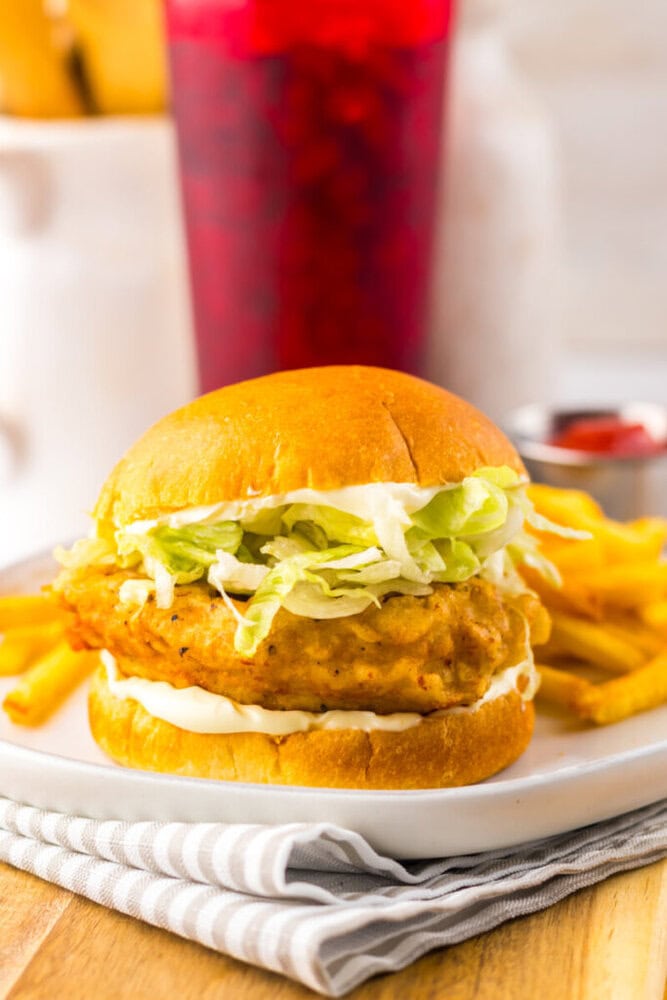 A fried chicken sandwich with lettuce and mayonnaise on a bun, served with French fries on a white plate. A red drink and ketchup are in the background.